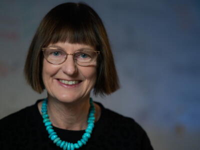 A photo of a woman with short, brown hair wearing glasses, a blue necklace and a black jumper. 